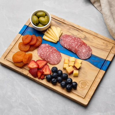 Wooden resin cutting board with assorted meats, cheeses, fruits, and olives on a light gray surface.