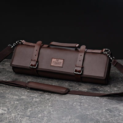 ITAMAE Yokohama Knifebag in dark brown premium leather with secure buckles, handle, and adjustable shoulder strap on a stone surface.