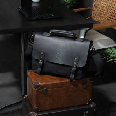 ITAMAE Black Out Messenger Bag placed on travel luggage – durable black leather messenger bag with adjustable shoulder strap.