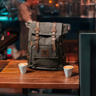ITAMAE Trooper Army Bag – waxed canvas and full-grain leather backpack for travel and everyday carry, shown in a coffee shop setting.